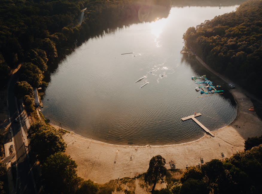 An aerial view of Ruislip Lido, featuring a large, irregularly shaped artificial lake surrounded by lush green grass, dense clusters of trees, and well-maintained walking paths. In the foreground, the